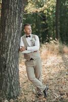 Portrait of the groom. The groom in a light gray suit is standing in the forest, leaning against a tree and looking away. Wedding in nature photo