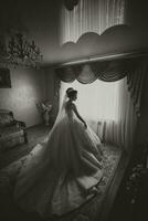 el novia en un blanco Boda vestido. contento hermosa joven mujer en blanco tradicional Boda vestir con largo tren en su habitación. grande ventanas y ligero paredes negro y blanco foto