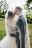 A curly-haired brunette bride in a white dress and a veiled groom embrace and kiss. Portrait of the bride and groom. Beautiful makeup and hair. Wedding in nature photo