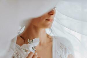 A brunette bride in a white dress holds her wedding bouquet, poses. French manicure. Open shoulders. Beautiful hands. Long veil. Morning of the bride. Details photo