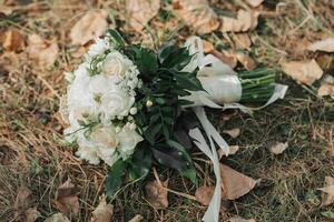 A wedding bouquet of white roses and greenery, decorated with a silk ribbon, lying on a background of dry leaves and grass. photo