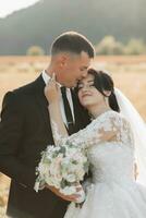 el novio en un negro traje y el novia son en pie en un campo en contra el antecedentes de montañas, el novio abrazos su amado desde detrás y Besos su. retrato. verano Boda en un brillante camino foto