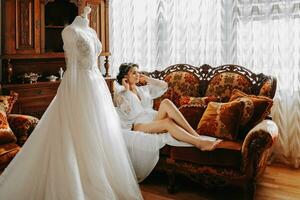 beautiful bride on a retro sofa in a robe with open legs, next to her dress, full-length photo. Wedding hairstyle, light makeup photo