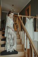 A delicate wedding dress hangs on a beautiful railing in the hallway of the house. The bride climbs the steps in a delicate transparent petticoat. Nice light. Wedding photo. A high quality photo