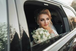 A blonde bride, smiling sincerely, sits in a black car on her wedding day with a bouquet of white roses. Portrait of the bride. Beautiful curls. Beautiful makeup and hair. A luxurious white dress photo