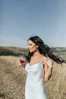 A girl with long black curly hair blowing in the wind is standing in a white silk dress. He holds a glass of champagne in one hand and touches his hair with the other. Portrait photo