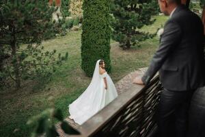 Wedding photo. The bride in a voluminous white dress and a long veil walks in the garden along a stone path, the groom stands on the balcony in the foreground, his shoulders turned to her. photo