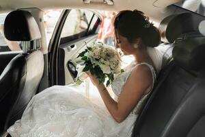 A young bride sniffs her wedding bouquet of white flowers while sitting in the car. Wedding photo. Summer wedding photo