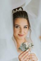 A brunette bride in a white dress holds her wedding bouquet, poses. French manicure. Open shoulders. Beautiful hands. Long veil. Morning of the bride. Details photo