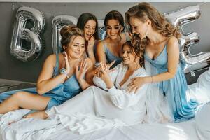 The bridesmaids look at the smiling bride. The bride and her cheerful friends are celebrating a bachelorette party on the bed in identical dresses. Bride and friends in the room photo