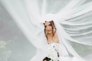 Curly brunette bride in a white dress, covered with a veil, poses for the camera with a bouquet of roses. Portrait of the bride. Beautiful makeup and hair. Wedding in nature photo