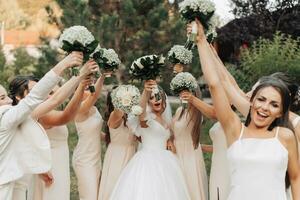 Wedding photo in nature. A brunette bride in a white long dress and her friends in nude dresses are standing against the background of trees, smiling, holding up their gypsophila bouquets. Young women