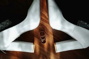 Photo of details at the wedding. Wedding rings of the bride and groom stand on white classic shoes, which are located on a wooden background. Contour line. Shadows and light.