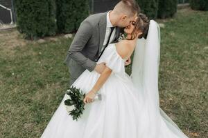 Portrait of the bride and groom in nature. The groom and the brunette bride in a white long dress are standing, kissing against the background of conifers. Stylish groom. Beautiful hair and makeup photo