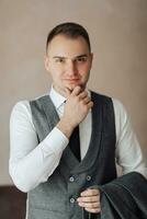 A man in a white shirt and a gray vest is posing by the window and looking at the camera, holding his jacket. Front view. A stylish watch. Men's style. Fashion. Business photo