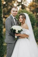 retrato de el novia y novio en naturaleza. recortado foto. el novia y novio pose, abrazando y sonriente, en contra el antecedentes de coníferas el novia en un largo vestir con un ramo de flores de rosas. foto