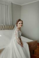 preparación para el boda. hermosa joven novia en blanco Boda vestir adentro. lujo modelo mirando fuera ventana a hogar en estudio habitación con grande ventana. el niña muestra foto