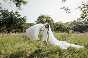 contento novia en blanco túnica bailando en jardín con Boda vestido. un velo ese fácilmente caídas en el verde césped. Mañana de el novia en el jardín. rústico estilo. primavera boda. brillante luz foto