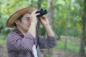 Asian man botanist holds binuculars to explore nature in forest. Concept, nature exploration. Ecology study. Pastime activity, lifestyle. Explore environment. Traveling alone. photo