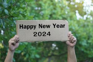 Close up hands hold paper card with text Happy New Year 2024. Outdoor background. Concept, greeting card for welcoming new year. photo