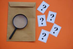 Brown envelope,magnifying glass and paper cards of question marks,orange background. Concept. Teaching aid. Education materials for doing activity or playing investigation games about find answers. photo