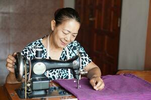 Happy senior Asian woman tailor is sewing cloth by vintage sewing machine at home. Concept, Senior lifestyle, spend free time to repair or design clothes. Hobby, not lonely. photo