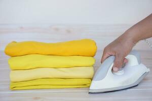 apilar de doblada amarillo ropa y mano sostiene eléctrico hierro en mesa. concepto, diario faena, familiar. planchado y doblada ropa para ordenado y limpio. mantenimiento y mantener vestidos. foto