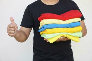 Close up man holds stack of colorful folded clothes after doing laundry. Thumbs up. Concept, daily chore, household. Folded clothes for neat and clean. Maintenance and keep garments for sanitary. photo