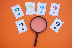 Paper cards of colorful question marks and magnifying glass on orange background. Concept. Teaching aid. Education materials for doing activity or playing investigation games about finding answers. photo