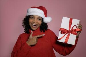 Cute smiling santa girl in red sweater and santa hat pointing at christmas gift box.winter holidays concept photo