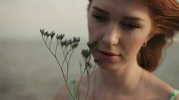 Portrait. Slow motion. Beautiful happy girl with red hair in a birch dress sits on the rocks with a flower on the sea background video