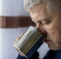 Portrait of the mid aged man with grey hair, wearing warm, dark blue sweater, drinking coffee. People photo