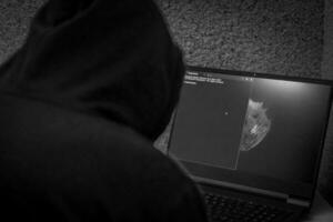 Man looked like hacker in a hood sitting in the front of laptop, writing a code. Cyber security photo