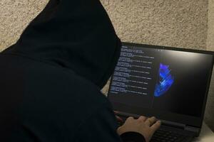 Man looked like hacker in a hood sitting in the front of laptop, writing a code. Cyber security photo