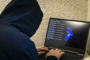 Man looked like hacker in a hood sitting in the front of laptop, writing a code. Cyber security photo