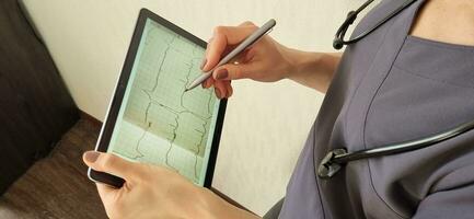 Shot of the female doctor in uniform against the white wall studying cardiogram on the tablet. Healthcare photo