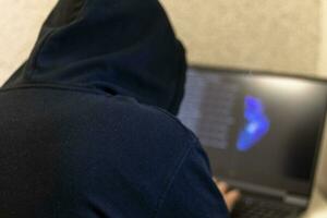 Man looked like hacker in a hood sitting in the front of laptop, writing a code. Cyber security photo