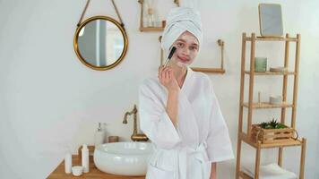 peau se soucier, après bain, produits de beauté à maison, caucasien femme, anti vieillissement. caucasien femme dans peignoir s'applique cosmétique masque à visage avec brosse tandis que permanent dans salle de bains video