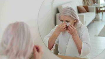 peau se soucier, Sénior femme, Accueil produits de beauté, personnes âgées âge, cosmétique procédures. personnes âgées femme 70 ans vieux les usages œil patchs tandis que séance dans de face de une miroir et souriant video