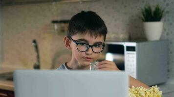 bleibe beim heim, Freizeit beim heim, Technologien zum Kinder, selbst Isolation. ein heiter Junge im Brille Uhren seine Liebling Cartoons während Sitzung beim Zuhause mit ein Laptop und Essen Popcorn. video