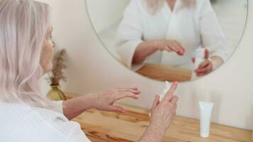 pelle cura, anziano donna, casa cosmetici, anziano età, cosmetico procedure. anziano donna usi mano crema mentre seduta nel davanti di un' specchio video