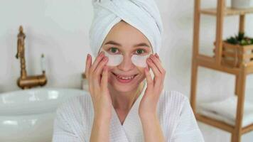 Skin Care, After Bath, Cosmetics at Home, Caucasian Woman, Anti Aging. Portrait.Caucasian woman in a robe and a towel on her head uses eye patches during cosmetic procedures looking at the camera video