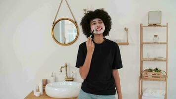 piel cuidado, productos cosméticos a hogar, natural crema, spa tratos, mezclado carrera. afro americano mujer aplicando máscara a cara con cepillo y sonriente mientras en pie en baño video