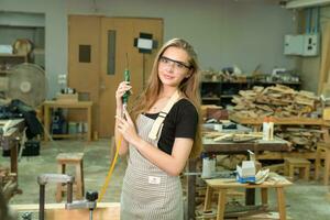 hembra carpintero vistiendo protector la seguridad lentes y utilizando eléctrico trabajo en un madera. artista o mueble diseñador trabajando en un producto idea en un taller. foto