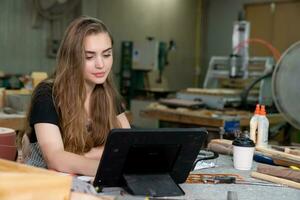 A young woman is training to be a carpenter in the workshop. She works with a laptop computer in a wood workshop. female carpenter contact customers by smartphone. SME orders, Start-up and small photo