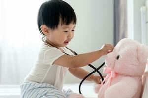 joven linda asiático niña jugando médico con favorito juguetes sentado en el cama. pequeño niña teniendo divertida. tomar cuidado osito de peluche oso jugando solo en el dormitorio, futuro, sueño, desarrollo, ef , iq, eq foto