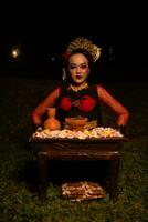 An Asian woman in a red dancer's dress is sitting at a ritual offering table full of flowers photo