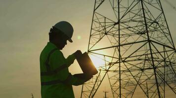 Engineer in standard safety uniform uses laptop to work, inspect high voltage pole with tablet near power pole video