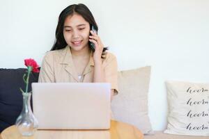 happy asian woman sitting on sofa answer call on smartphone and working with laptop wearing casual clothes, female designer working on project at home photo