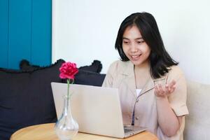 smiling asian woman working on laptop with raising palms sitting on couch wearing casual clothes, presenting gesture indoors photo